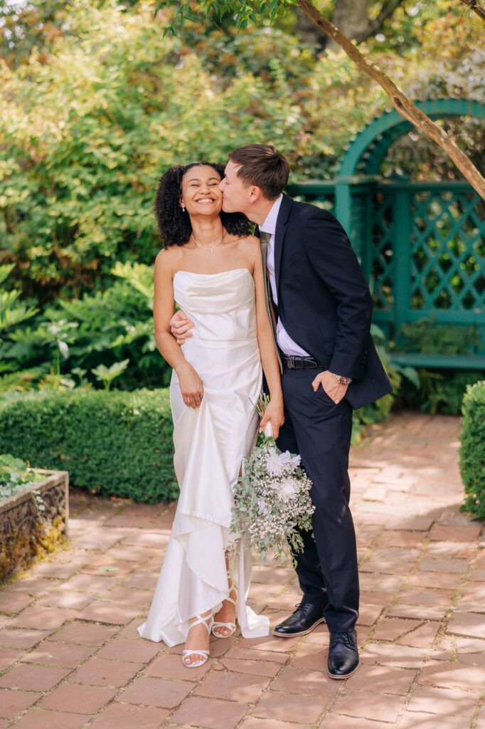 Groom kisses the bride on the cheek as she smiles joyfully, holding a soft lavender and greenery bouquet, during their garden wedding portrait session on a brick pathway surrounded by lush greenery and a teal arbor.