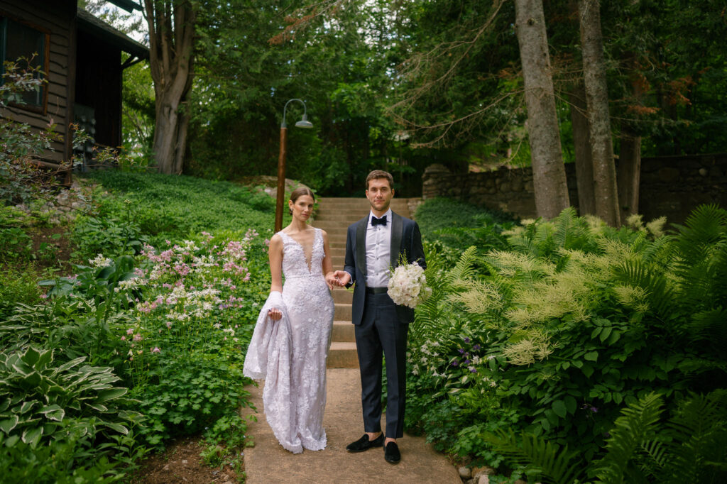 Bride and groom walk hand-in-hand down a garden path surrounded by lush summer greenery and blooming flowers at Whiteface Club in Lake Placid, New York; the bride wears a lace gown with a deep neckline, and the groom carries a white bouquet in a tailored tuxedo.