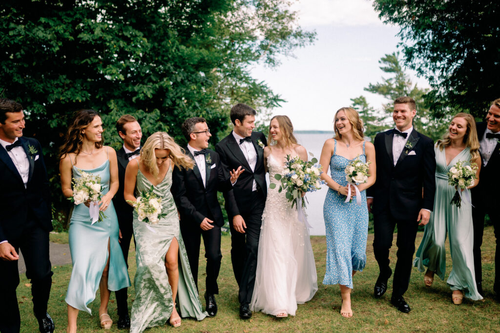 Bride and groom walk joyfully with their wedding party along the shoreline at a private estate on Lake Champlain, surrounded by friends in colorful dresses and tuxedos, all laughing and holding bouquets.