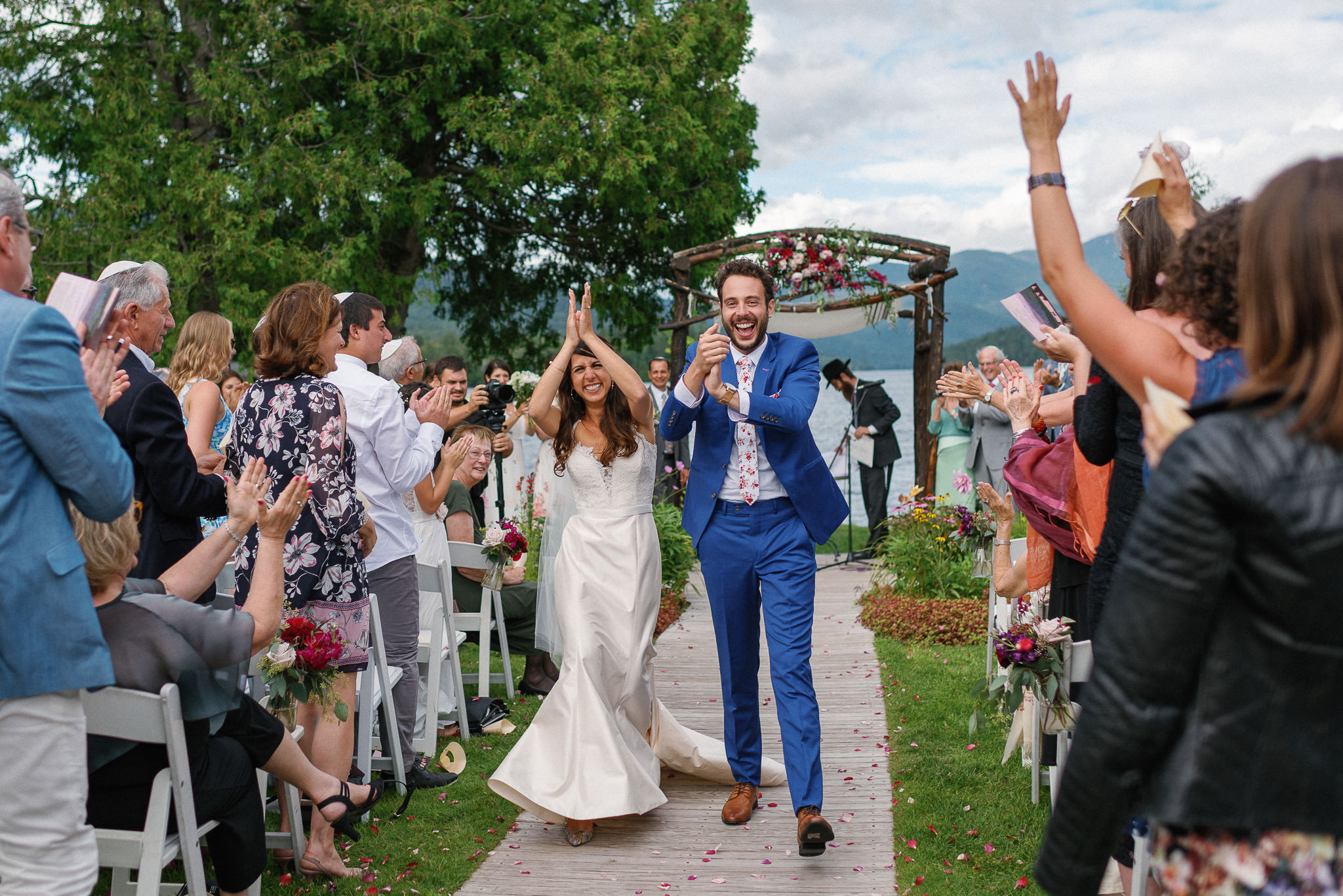 Bride and groom walk joyfully down the aisle as newlyweds at an outdoor lakeside ceremony, surrounded by cheering guests under a floral-adorned chuppah at Whiteface Club in Lake Placid, New York.