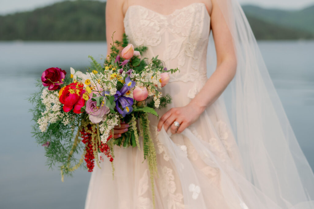 Close-up of a bride holding a vibrant spring bouquet with tulips, peonies, irises, and wildflowers, wearing a lace-embroidered strapless gown, standing lakeside at Whiteface Club in Lake Placid, New York.