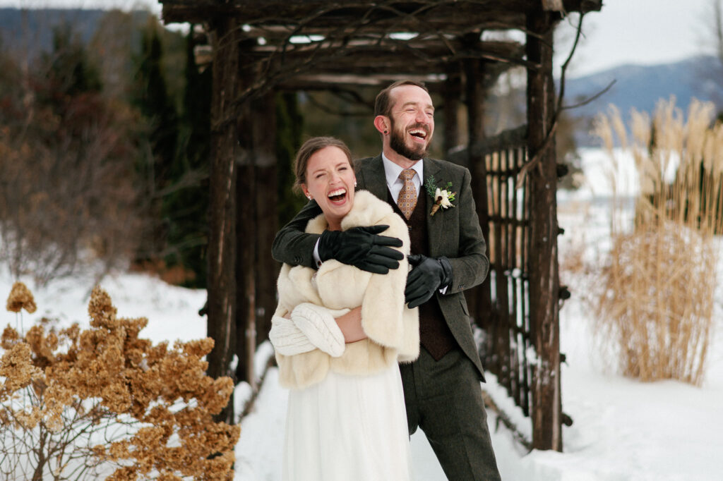 Bride and groom laugh joyfully while embracing in the snow beneath a rustic wooden arbor during their winter wedding at Whiteface Club in Lake Placid, New York.