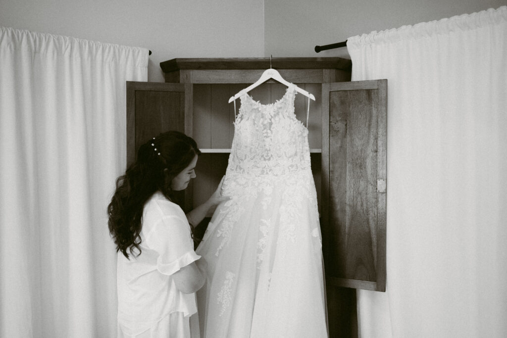 Bride looking lovingly at her lace wedding dress hanging in a wooden armoire, captured in a quiet getting-ready moment at Applegate Ranch.