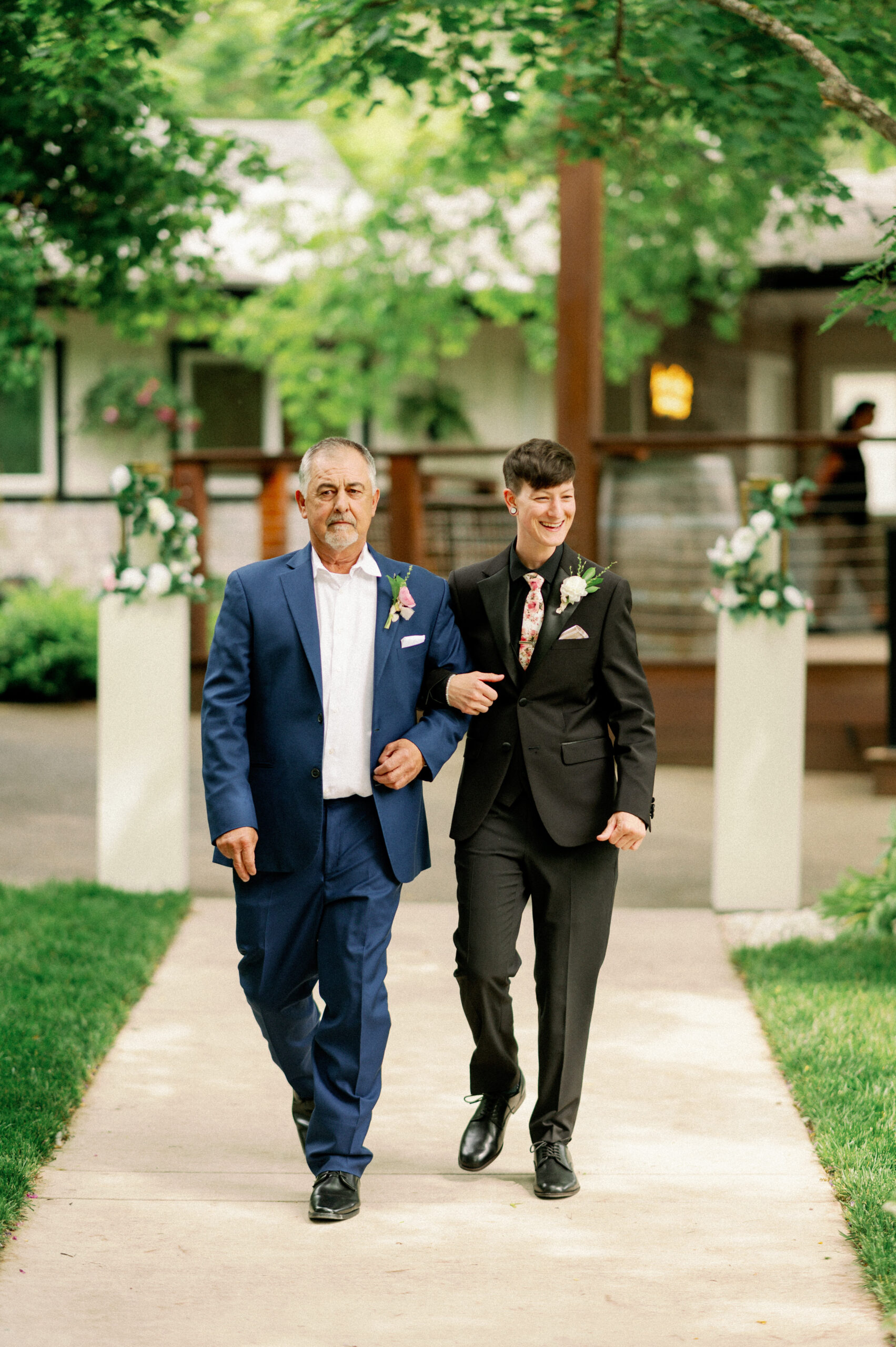 One bride, dressed in a black suit, walks arm-in-arm with a family member down the aisle surrounded by greenery at Applegate Ranch.