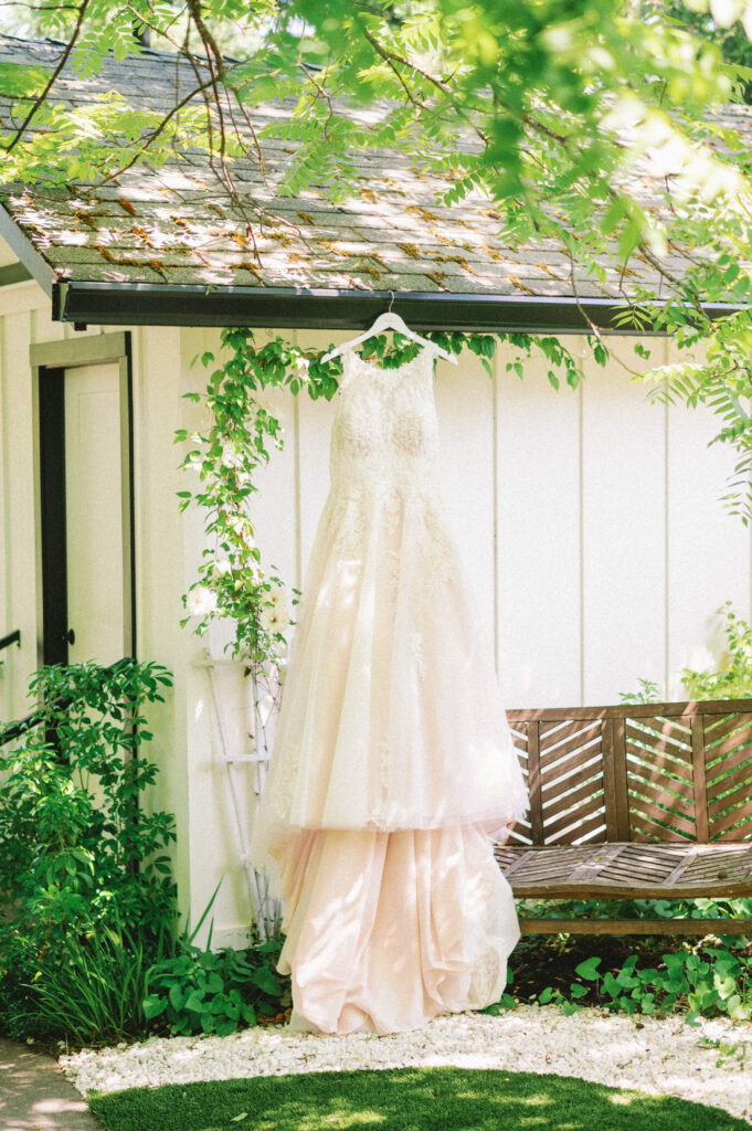Lace wedding gown hanging from a porch beam at Applegate Ranch, framed by dappled sunlight and greenery before the ceremony.