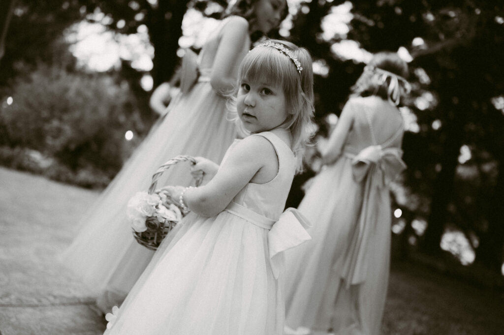 Young flower girl glances back over her shoulder while holding a flower basket, surrounded by bridesmaids in tulle dresses.