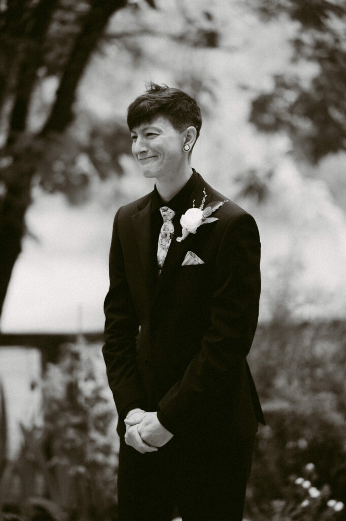 Jocelyn smiling during the ceremony, wearing a black suit with a floral tie and boutonniere, in front of blurred greenery.