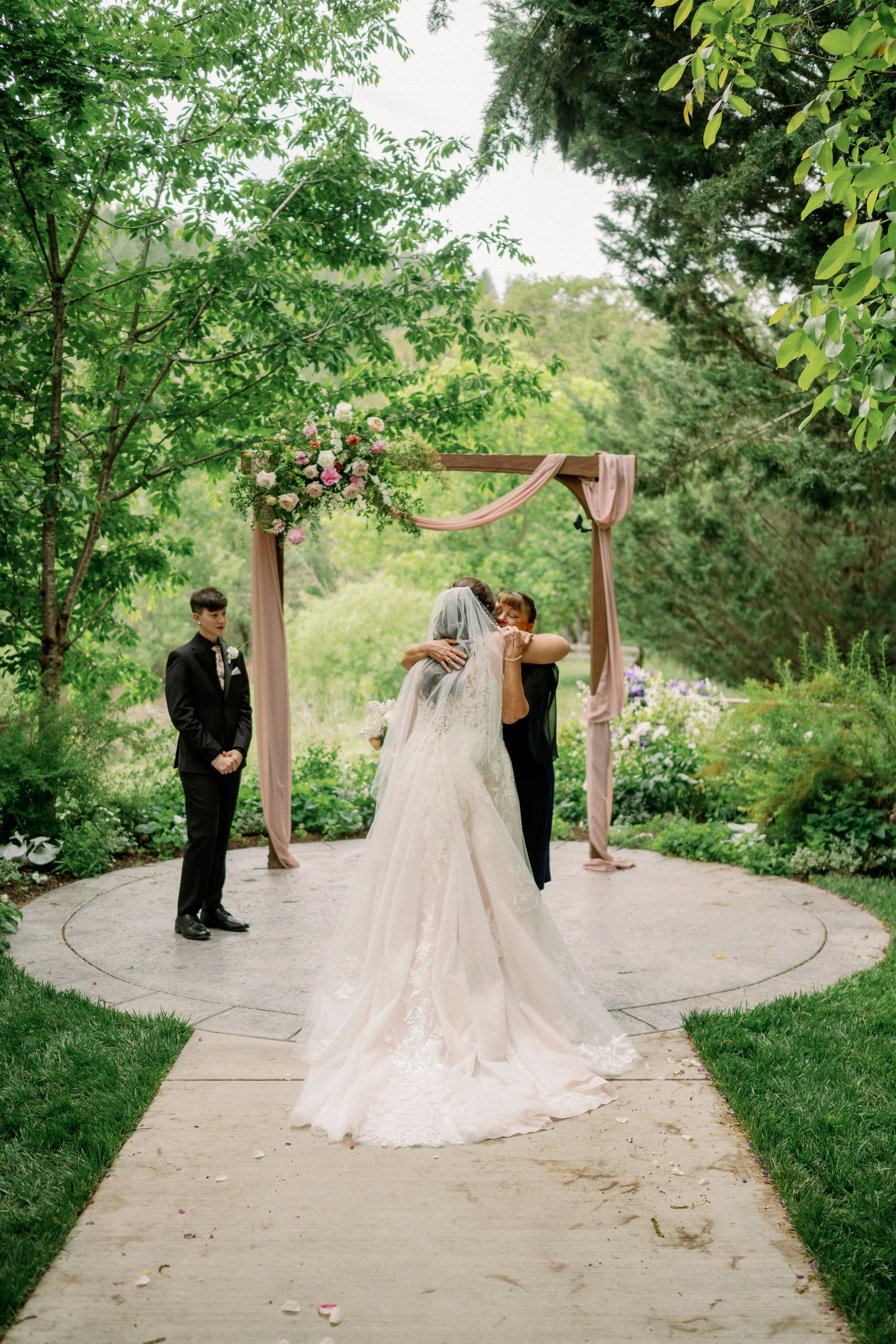 At the altar, Bailey hugs her mother while Jocelyn watches, standing beneath the floral wedding arch.