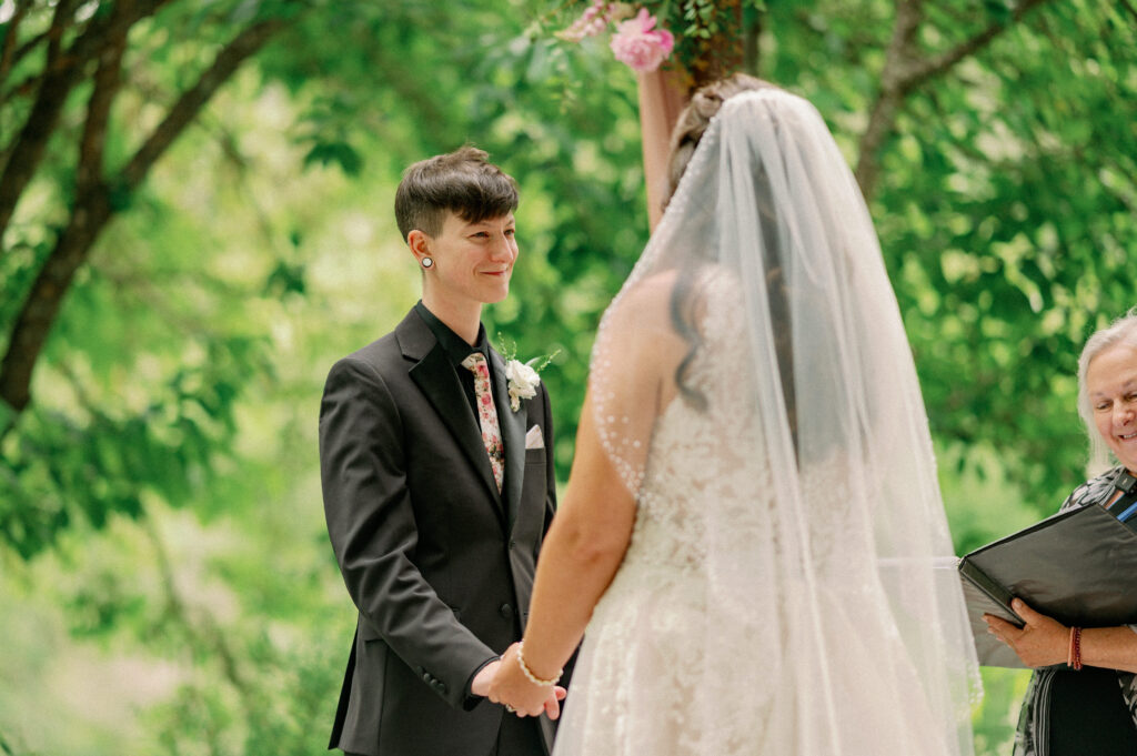 One bride smiles at her partner while holding hands during their ceremony, greenery and florals surrounding them.