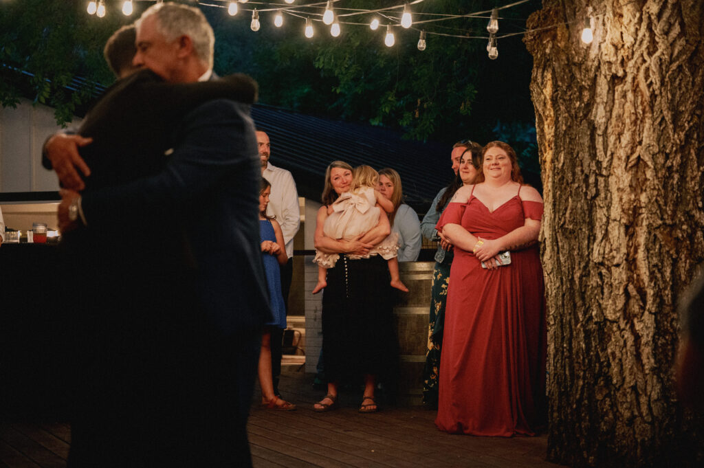 Guests gather on a warmly lit patio, watching an emotional embrace during a wedding reception dance.