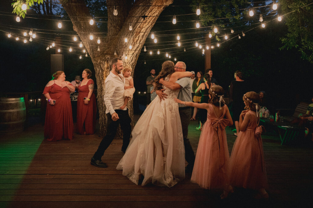 The bride shares a heartfelt hug with a guest under twinkle lights on the outdoor dance floor as family and friends look on.
