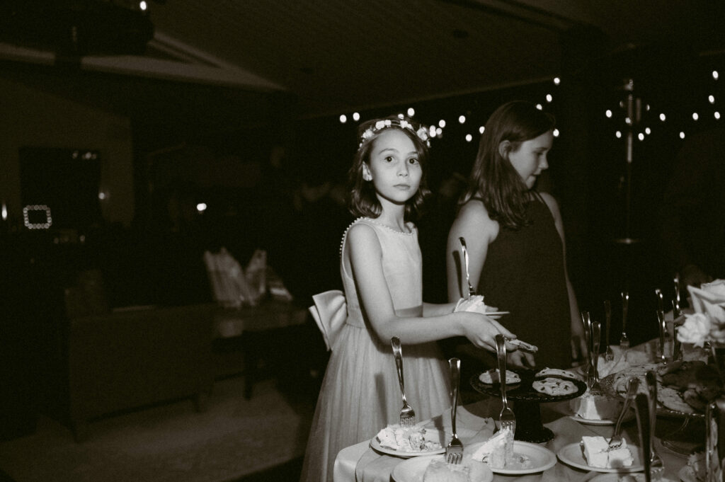 A flower girl in a tulle dress with a floral crown pauses mid-slice at the dessert table during the wedding reception.