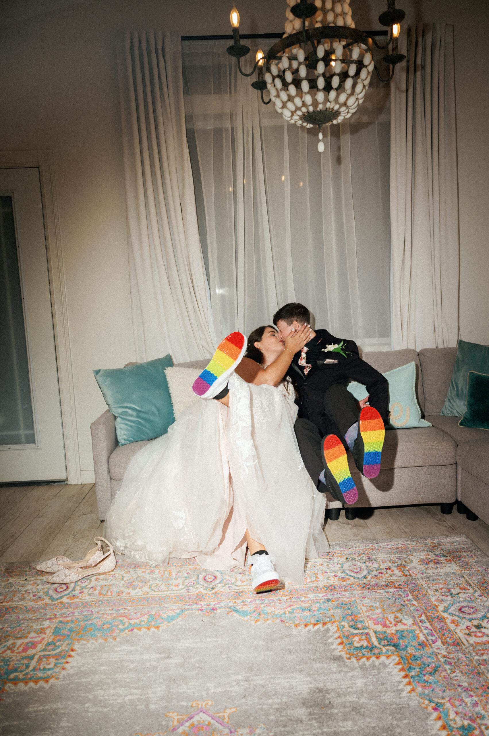 A LGBTQ couple sitting on a couch, sharing a kiss, with their colorful-soled shoes visible. One bride is wearing a white dress, and the other bride is in a black outfit. The setting features a chandelier and light-colored curtains.