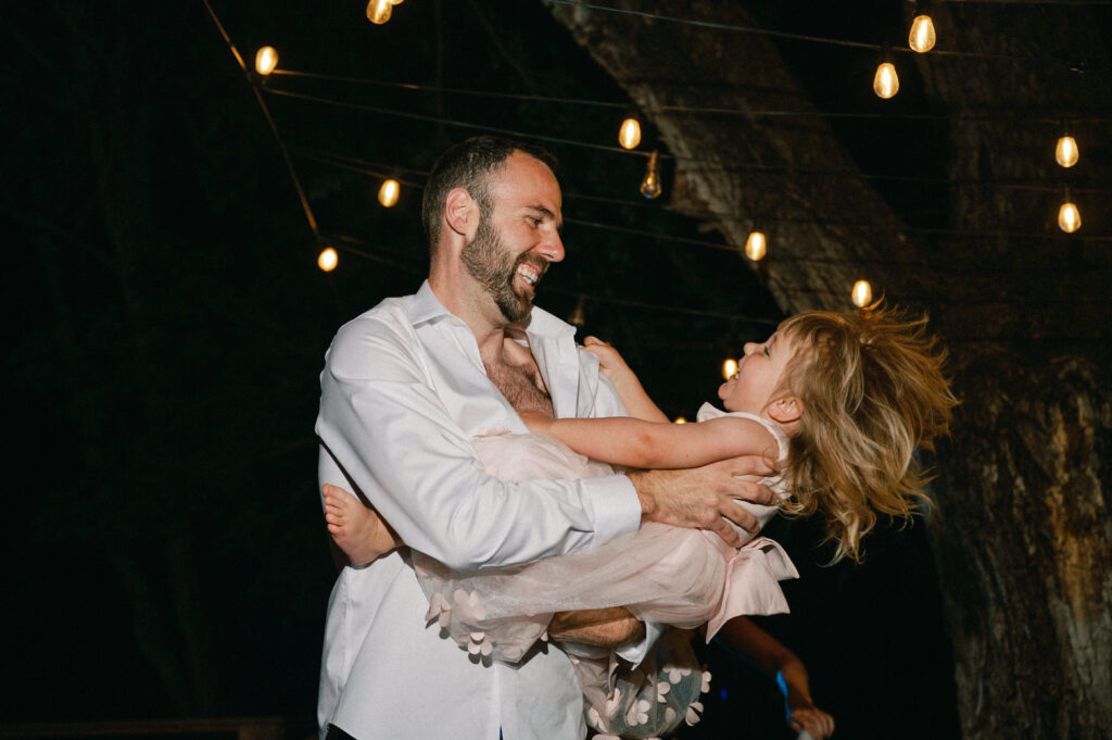 A father twirls his young daughter on the dance floor, both laughing under the string lights.