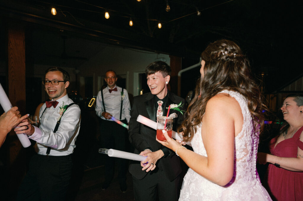 The newlyweds laughing and dancing with their wedding party, illuminated by warm overhead string lights.