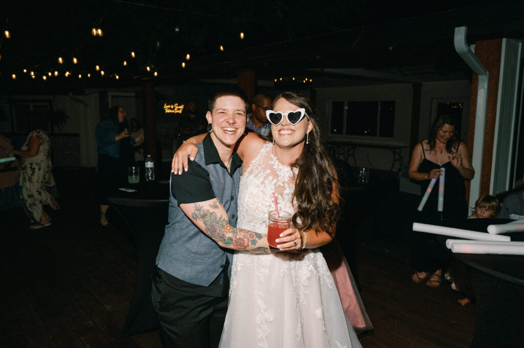The bride, wearing heart-shaped sunglasses, poses with a guest during the reception party, both smiling and holding drinks.