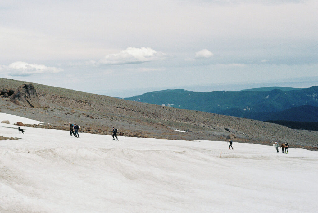 Skiers and hikers move across a late-season snowfield on Mount Hood, with wide mountain views stretching into the distance.