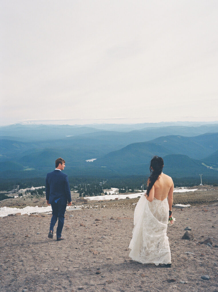 The bride and groom walk down the mountain path together, looking out over the sweeping forested landscape below Mount Hood.