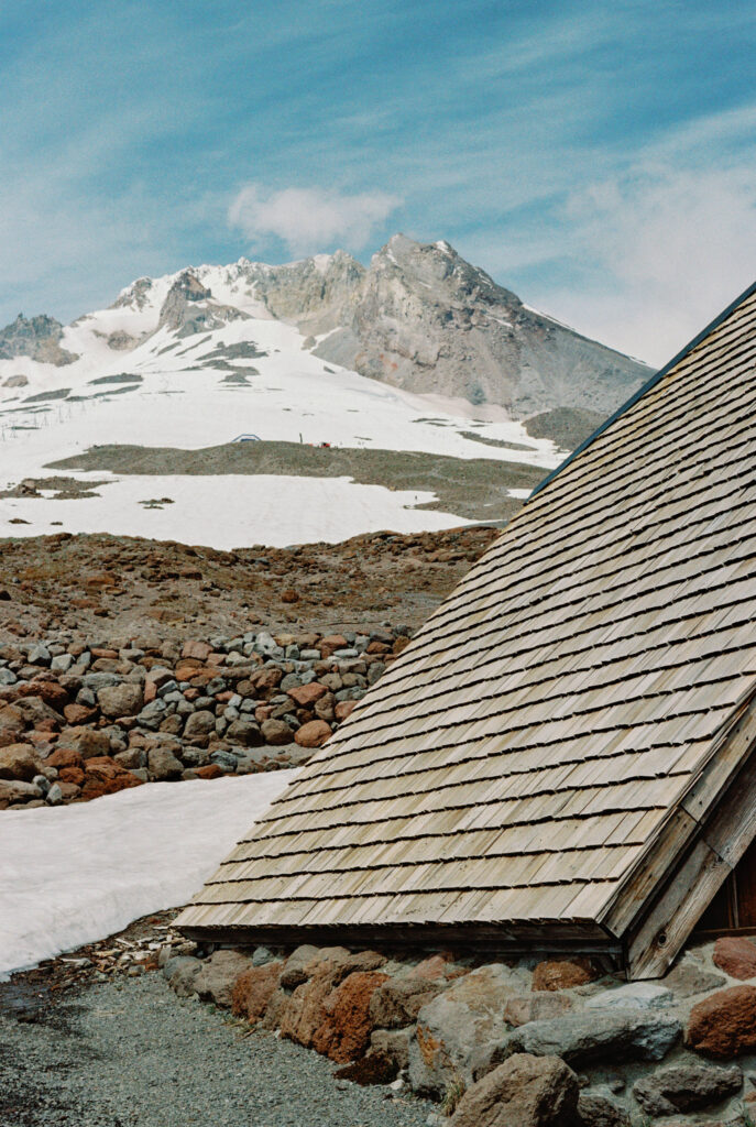 A view of Mount Hood’s snow-covered slopes rising behind the wooden roofline and stone foundation of Silcox Hut.