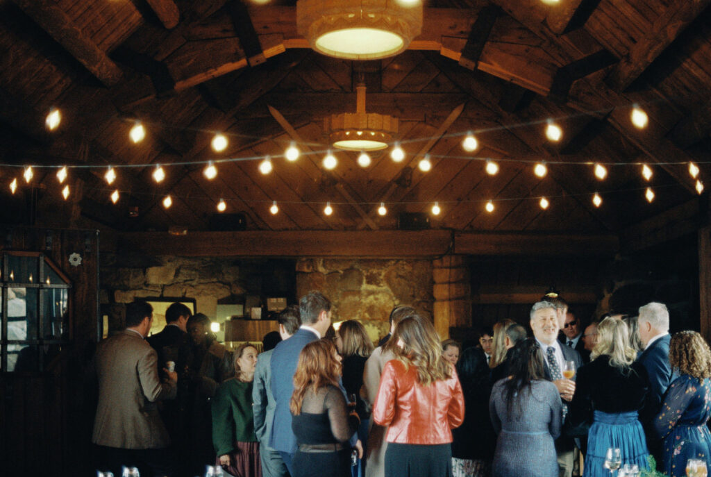 Wedding guests gather and mingle beneath warm string lights inside the rustic stone interior of Silcox Hut.
