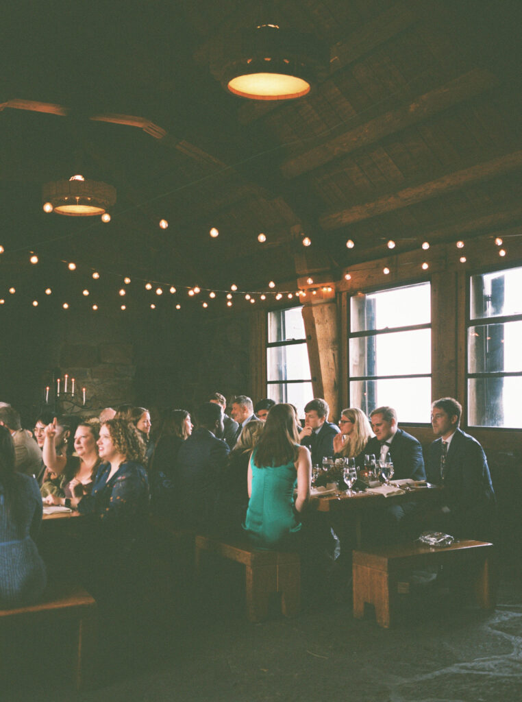 Wedding guests enjoy dinner beneath warm string lights inside the cozy, timber-and-stone interior of Silcox Hut.