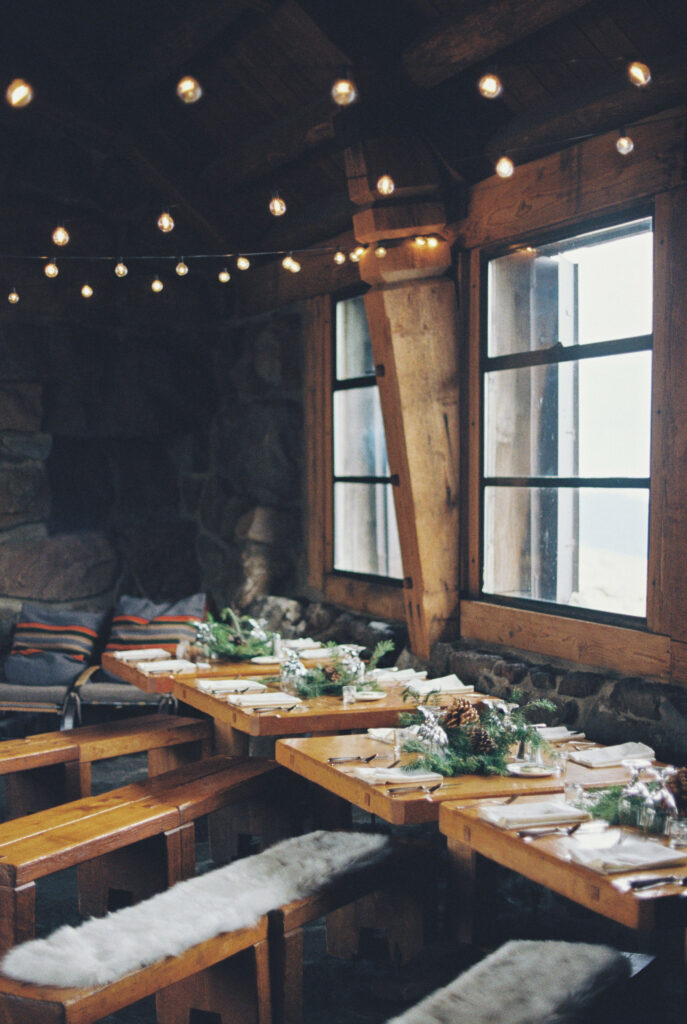 A film photograph of rustic wooden tables decorated with greenery under warm string lights inside Silcox Hut’s stone interior.