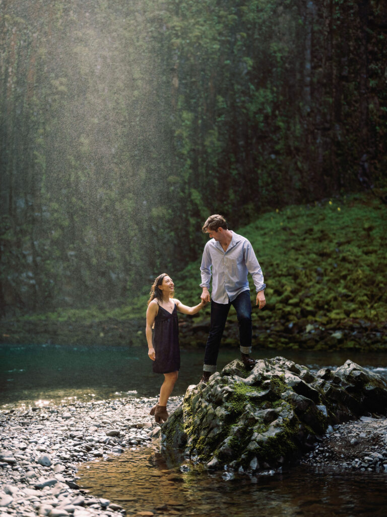 A man and woman smile as they climb on rocks at their engagement session at Abiqua Falls.