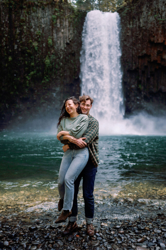 A man playfully hugs a woman from behind, lifting her in the air while Abiqua Falls roars in the background.