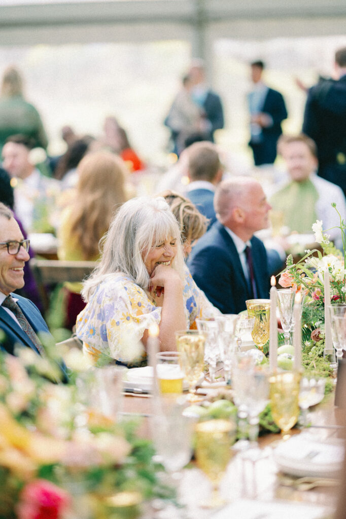 Wedding guests seated at long reception tables beneath a clear tent, surrounded by florals, candlelight, and soft summer light.