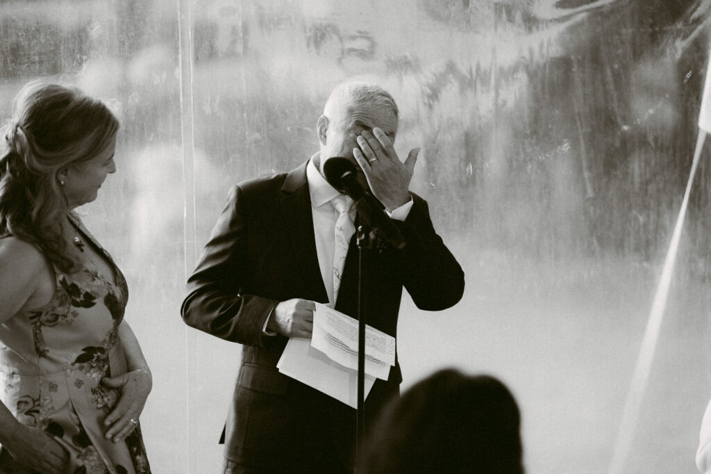 Black and white image of a father wiping away tears while giving a speech during the wedding reception.