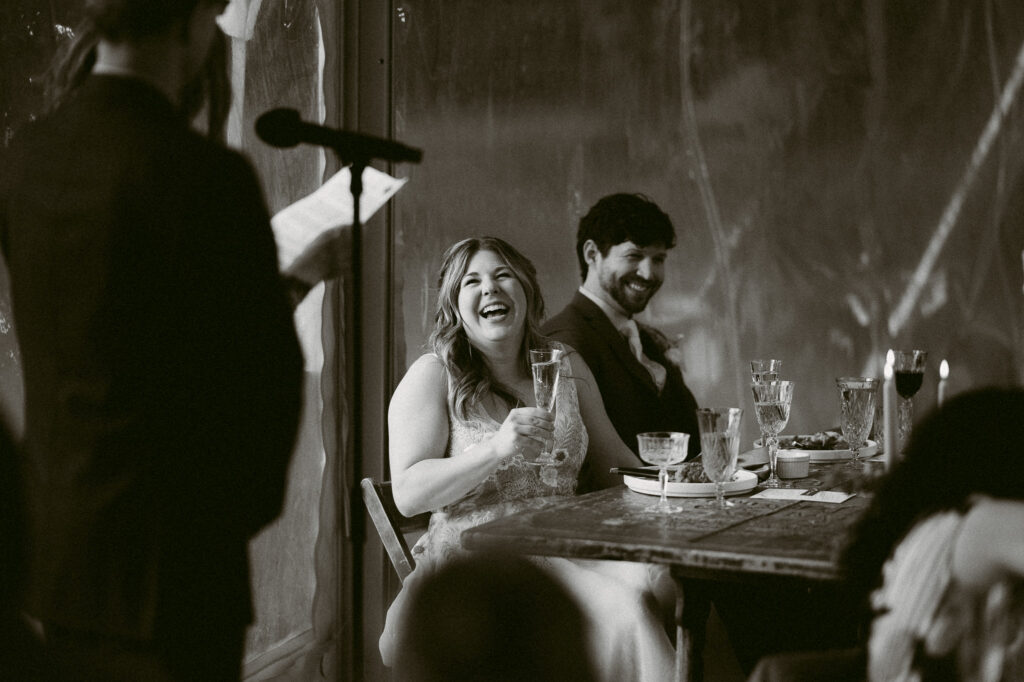 Black and white photo of the bride laughing during a toast at the reception, holding a champagne glass as the groom smiles beside her.