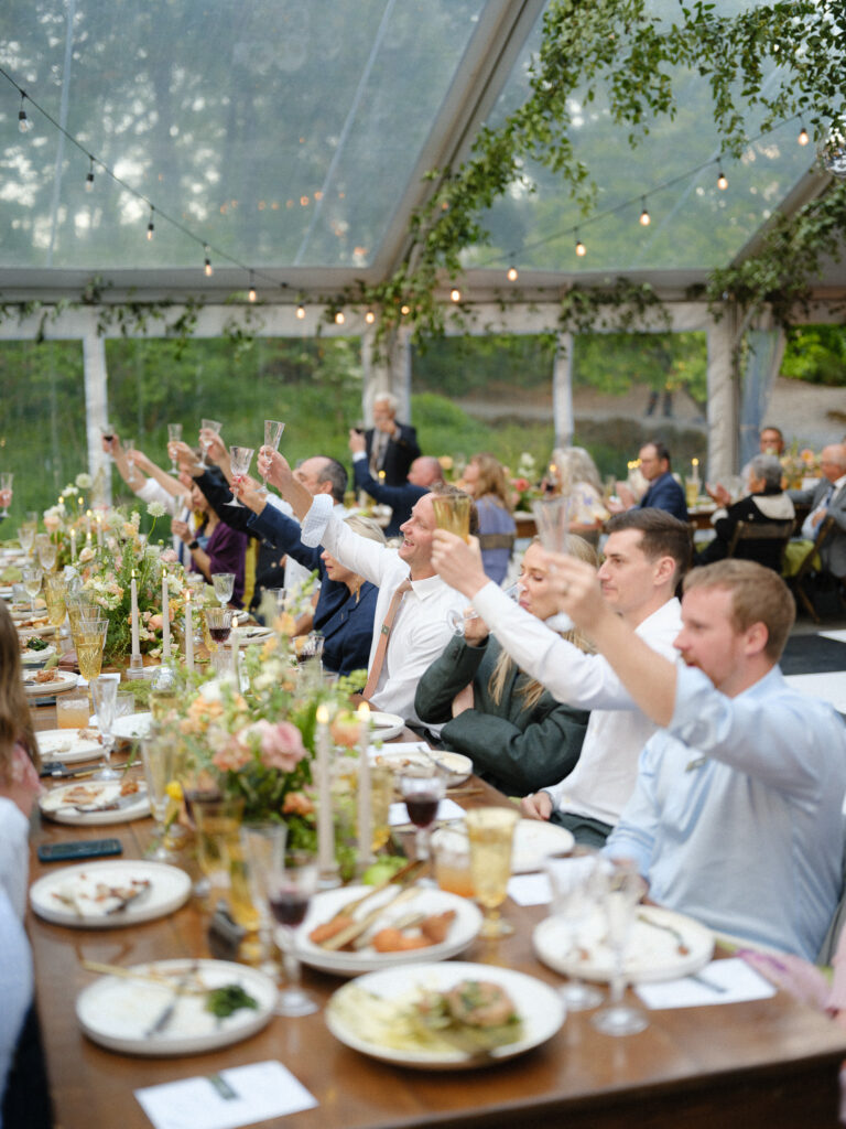 Wedding guests raising champagne glasses during a joyful toast at a long reception table beneath a clear tent at the Griffin House.