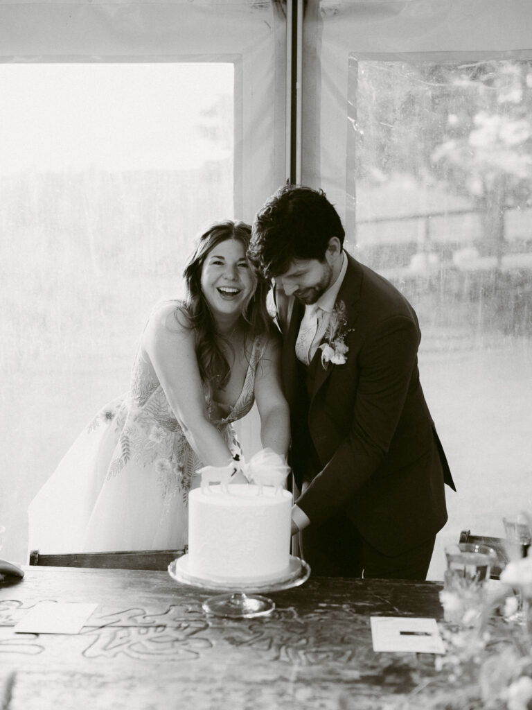 Black and white image of the couple laughing together while cutting their wedding cake during the reception