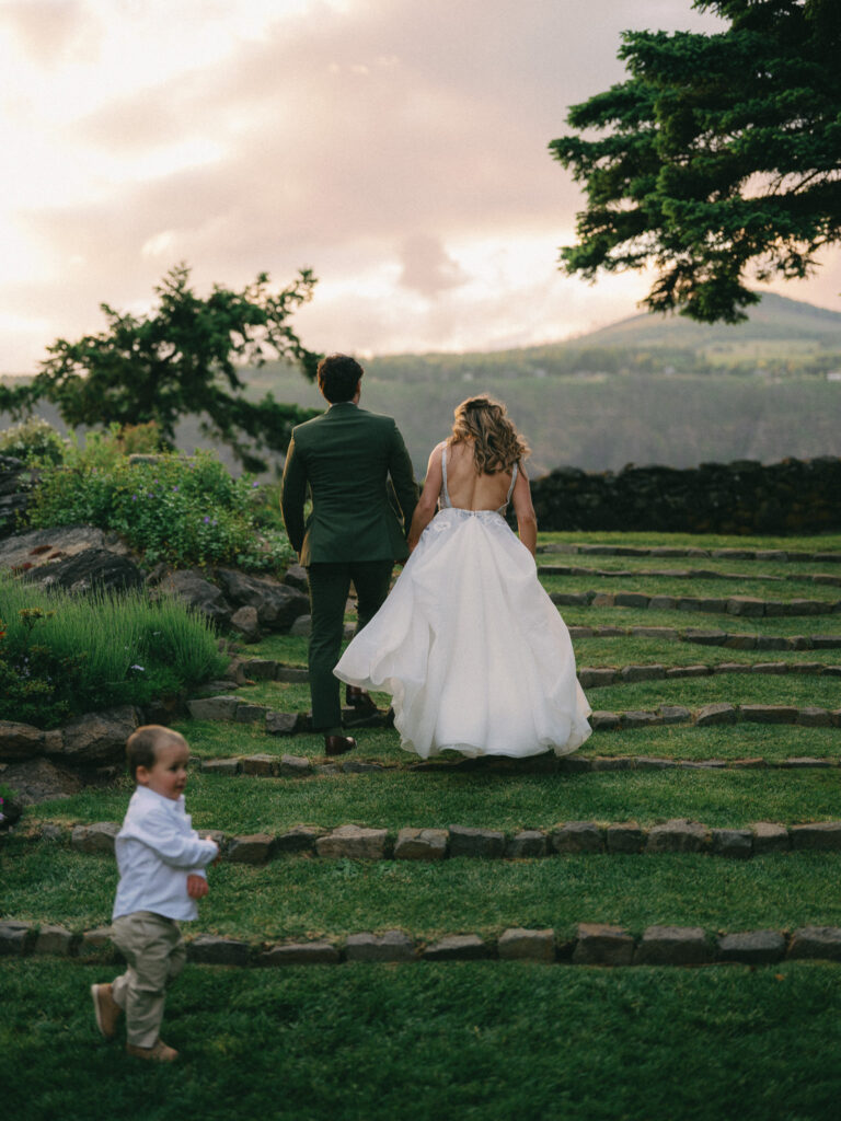 The newlyweds walking hand in hand across the Griffin House lawn at sunset, with stone steps and the Columbia River Gorge in the distance.