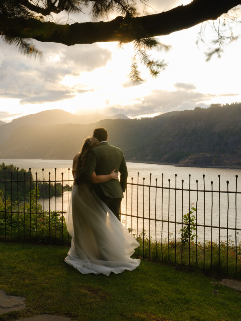 The couple standing together at sunset, overlooking the Columbia River Gorge from the Griffin House, framed by tree branches and warm evening light.