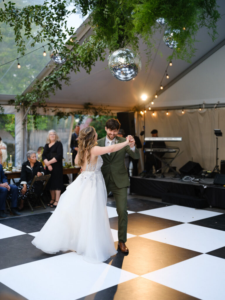 The couple sharing their first dance beneath a clear tent at the Griffin House, surrounded by hanging greenery, disco balls, and guests.