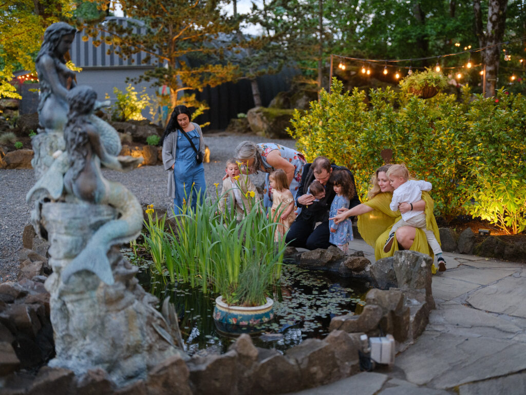 Wedding guests gathered around a small pond at the Griffin House, pointing and laughing as children explore the water during golden hour.