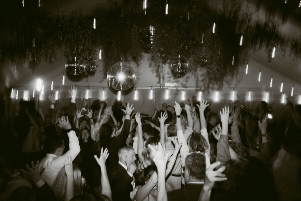 Wide black and white photo of a packed dance floor beneath a clear tent, with hands raised and disco balls overhead.