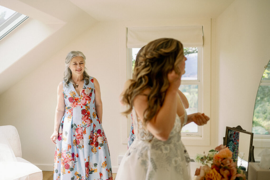 The mother of the bride smiling as she watches the bride adjust her dress during getting ready.
