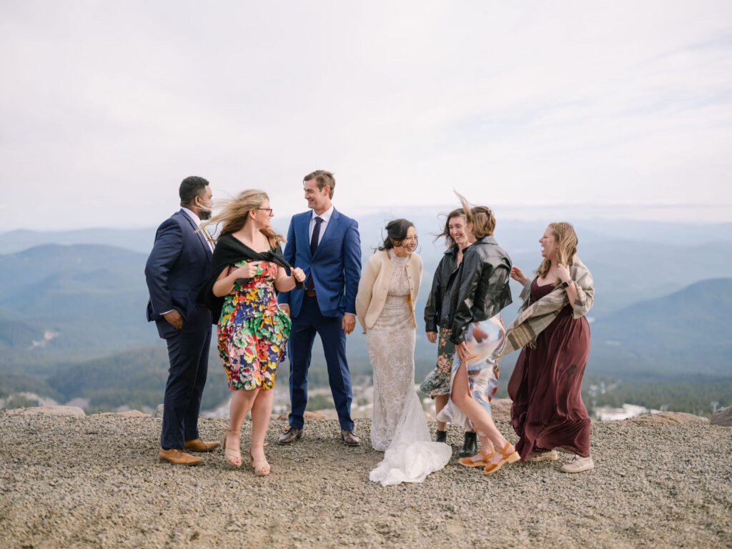 The couple and their wedding party laugh together on a windy mountain overlook above Timberline Lodge.