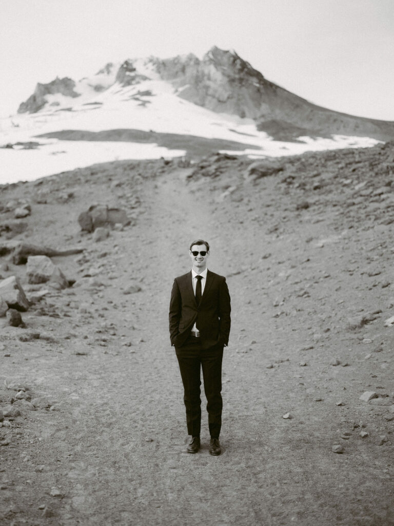 The groom stands on a rocky mountain path in front of the snow-covered slopes of Mount Hood during his Silcox Hut wedding portraits.