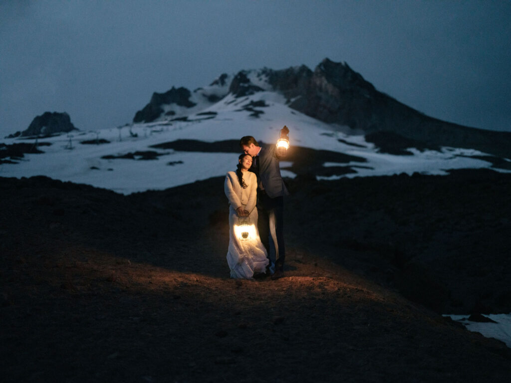 A bride and groom hold glowing lanterns at dusk on the snowy slopes of Mount Hood during their Silcox Hut wedding portraits.