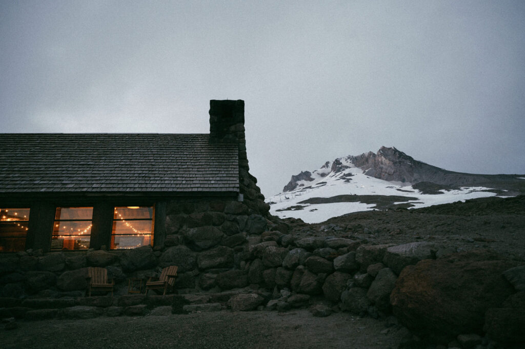 Silcox Hut glows with warm golden light at dusk as Mount Hood rises snow-covered behind the stone cabin.