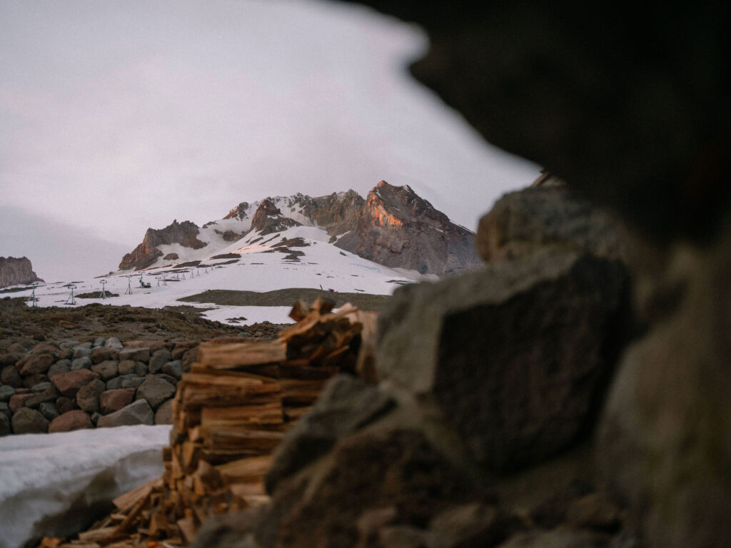 Early morning light hits the summit of Mount Hood, viewed between rocks and stacked firewood near Silcox Hut.