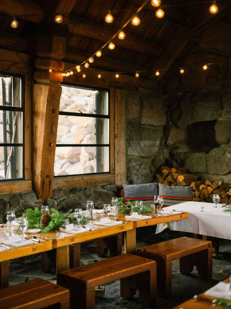 Rustic wooden tables decorated with pine greenery and glowing string lights inside the stone interior of Silcox Hut’s wedding reception space.