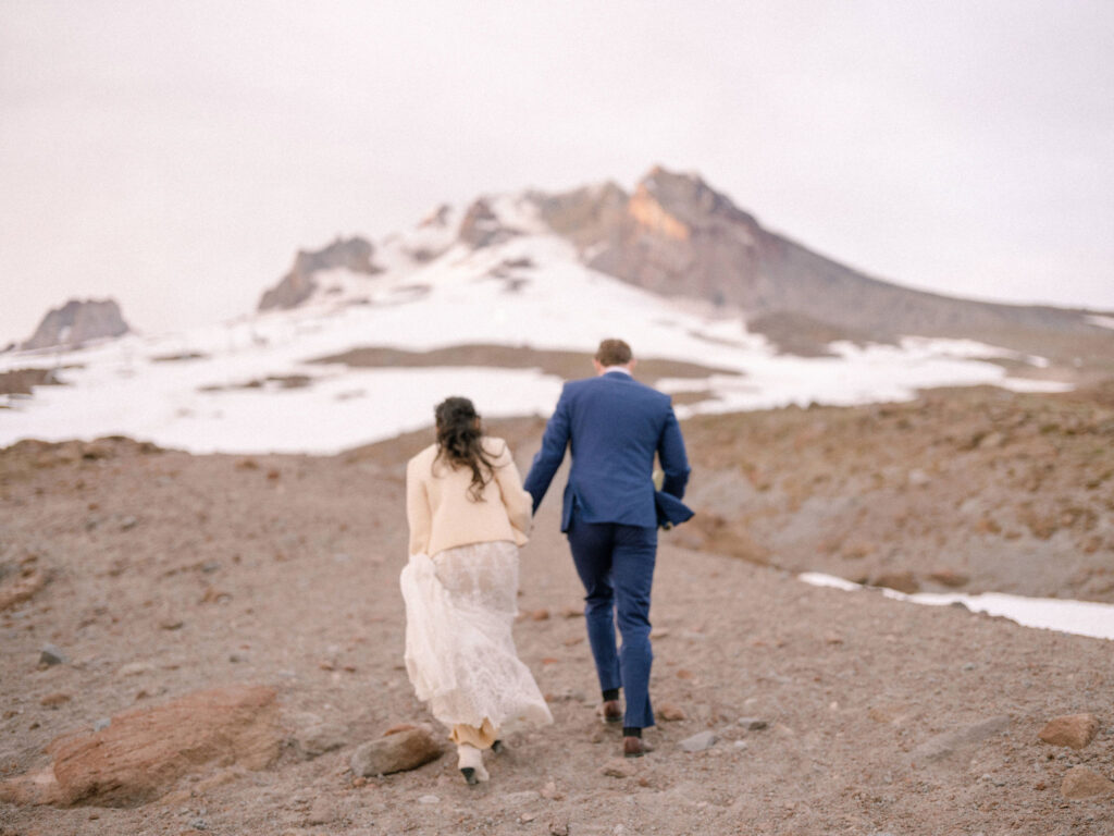 A bride and groom walk hand in hand up the rocky, snow-dusted slopes of Mount Hood during their early summer Silcox Hut wedding.