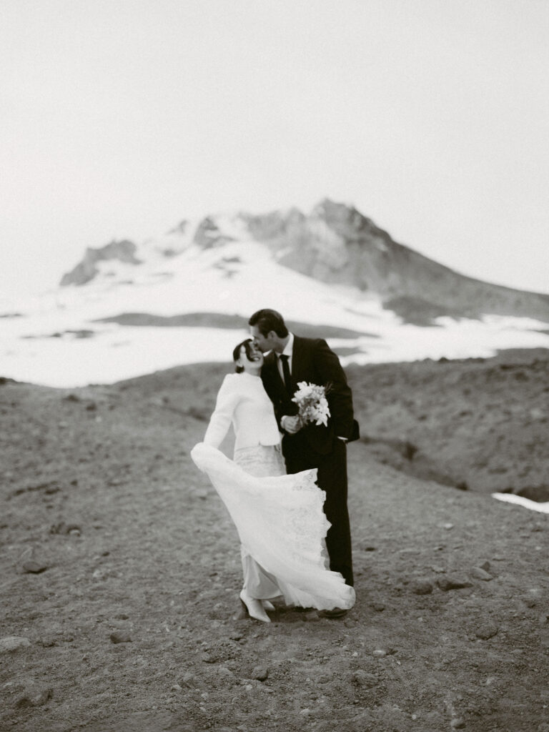 The bride and groom share a kiss on a windy mountainside as her lace dress flows in the air, with Mount Hood rising behind them.