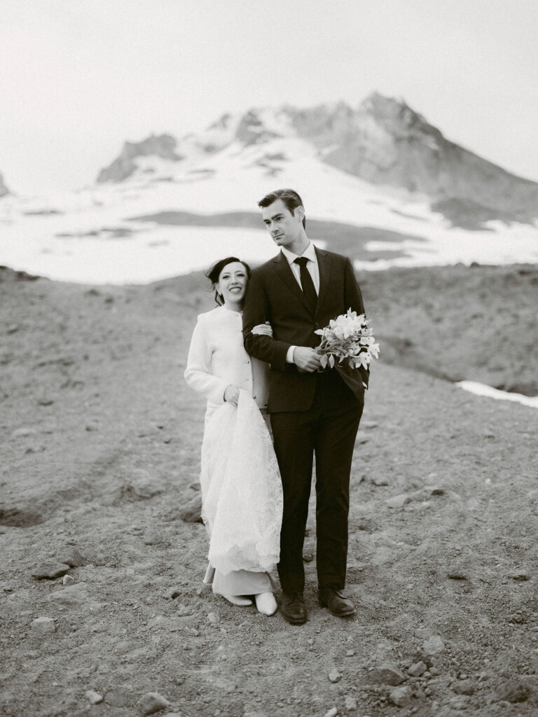 The bride and groom stand arm-in-arm on the mountain slope beneath Mount Hood, sharing a quiet moment during their Silcox Hut wedding portraits.