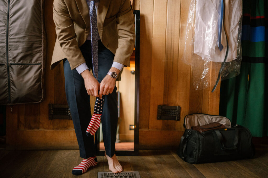 The groom puts on red, white, and blue socks while getting dressed inside Timberline Lodge before the Silcox Hut wedding.