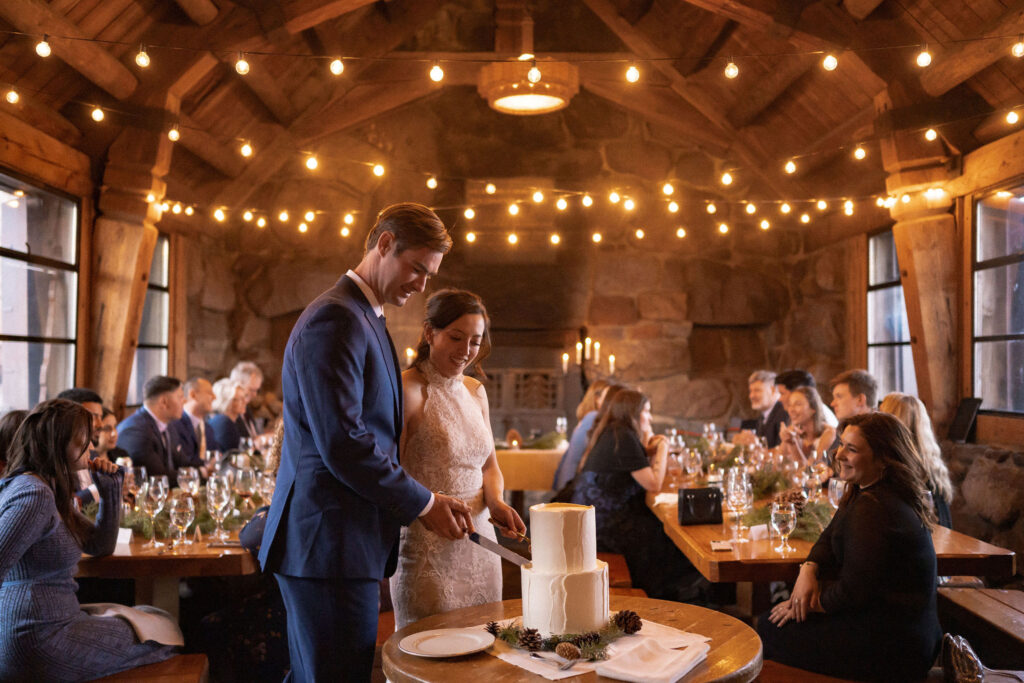 The bride and groom cut their wedding cake inside Silcox Hut, surrounded by guests and warm string lights in the rustic stone space.